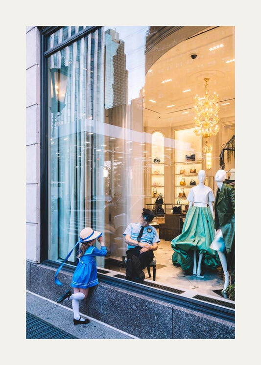 A street-style photograph of a boutique window display, featuring a young girl in a blue sailor-style dress and hat leaning against the glass, with a sleeping man in a uniform inside the shop and mannequins wearing a white top and green skirt.