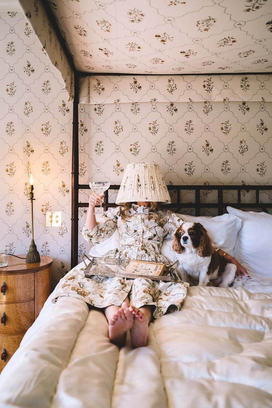 Blending In - Nick Mele Print of a women laying on a fancy canopy bed with a martini in her hand. Her dress matches the wall paper and she has a matching lamp shade over here head. There is a little dog next the her on the bed. 