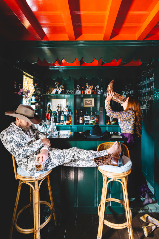 Man and woman at a bar. woman in purple dress drinking from the man's boot while the gentleman sleeps in a barstool.