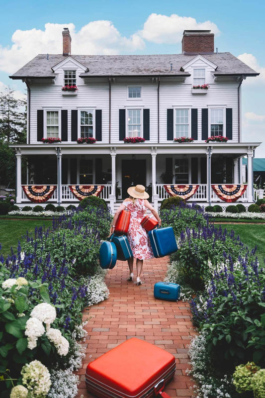 Nick Mele Fine Art Print, titled Checking In - Woman with suitcases walking towards a large white house with a garden.