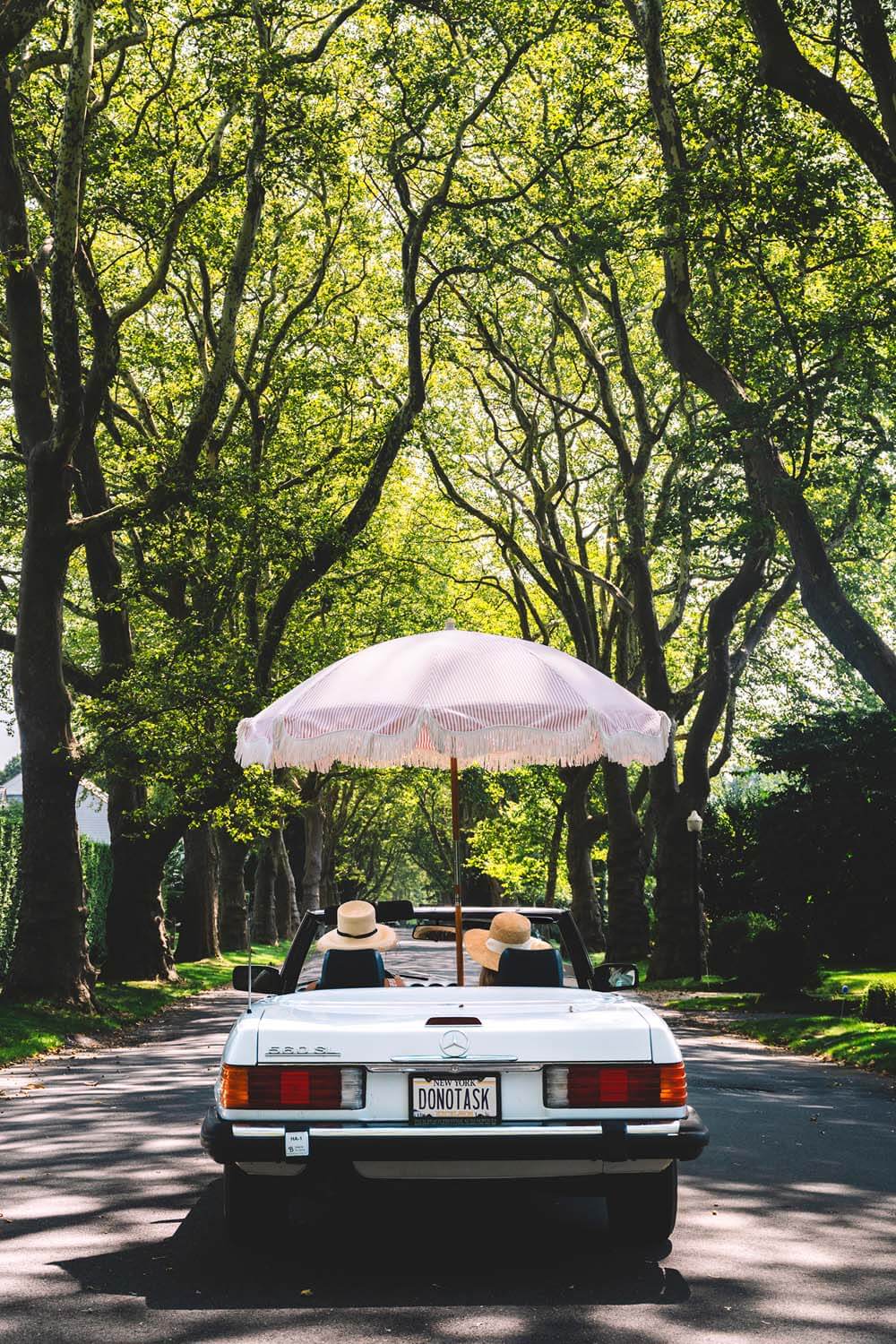 Nick Mele print titled "Cruisin" - A white convertible car with a pink umbrella in the convertable on a tree-lined road