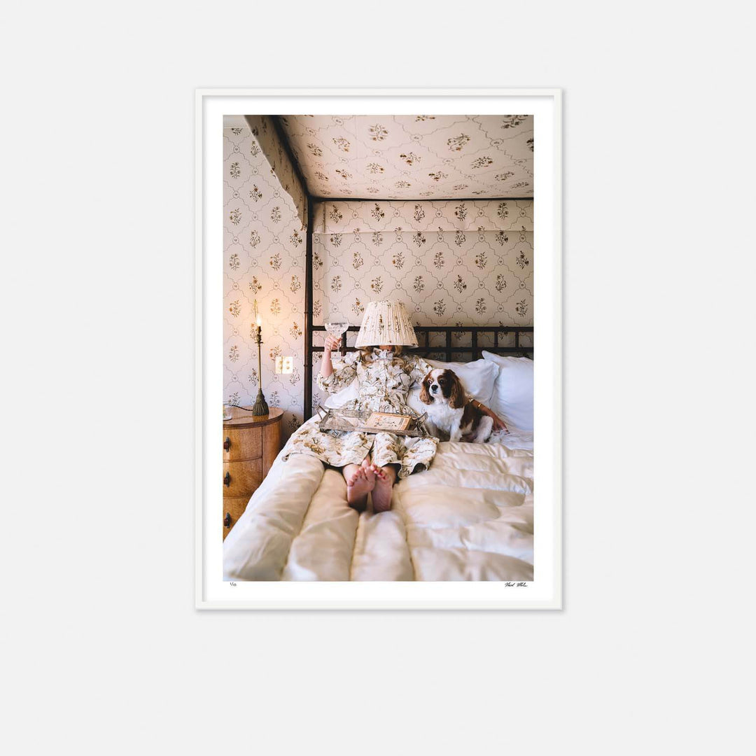 Nick Mele Framed Print - Blending In - Person lying on a bed with a dog, surrounded by floral wallpaper and a lamp.