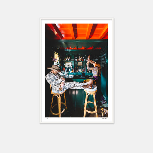 Man and woman at a bar. woman in purple dress drinking from the man's boot while the gentleman sleeps in a barstool.