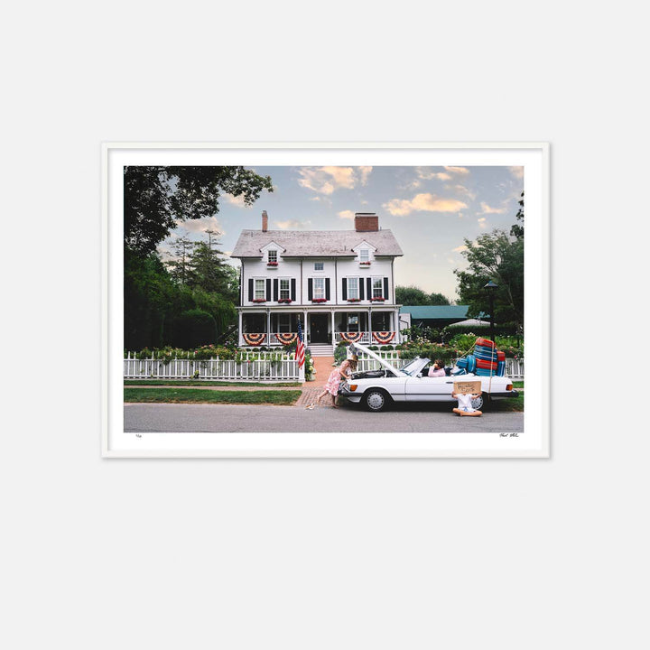 Nick Mele Print titled "Heading south". A white house with American flags, a man sitting ind river seat of convertable. Tons of lugage strapped to back of car. A women is leaning into hood of car trying to fixe it and a little boy is sitting outside car with a sign that says Heading south.