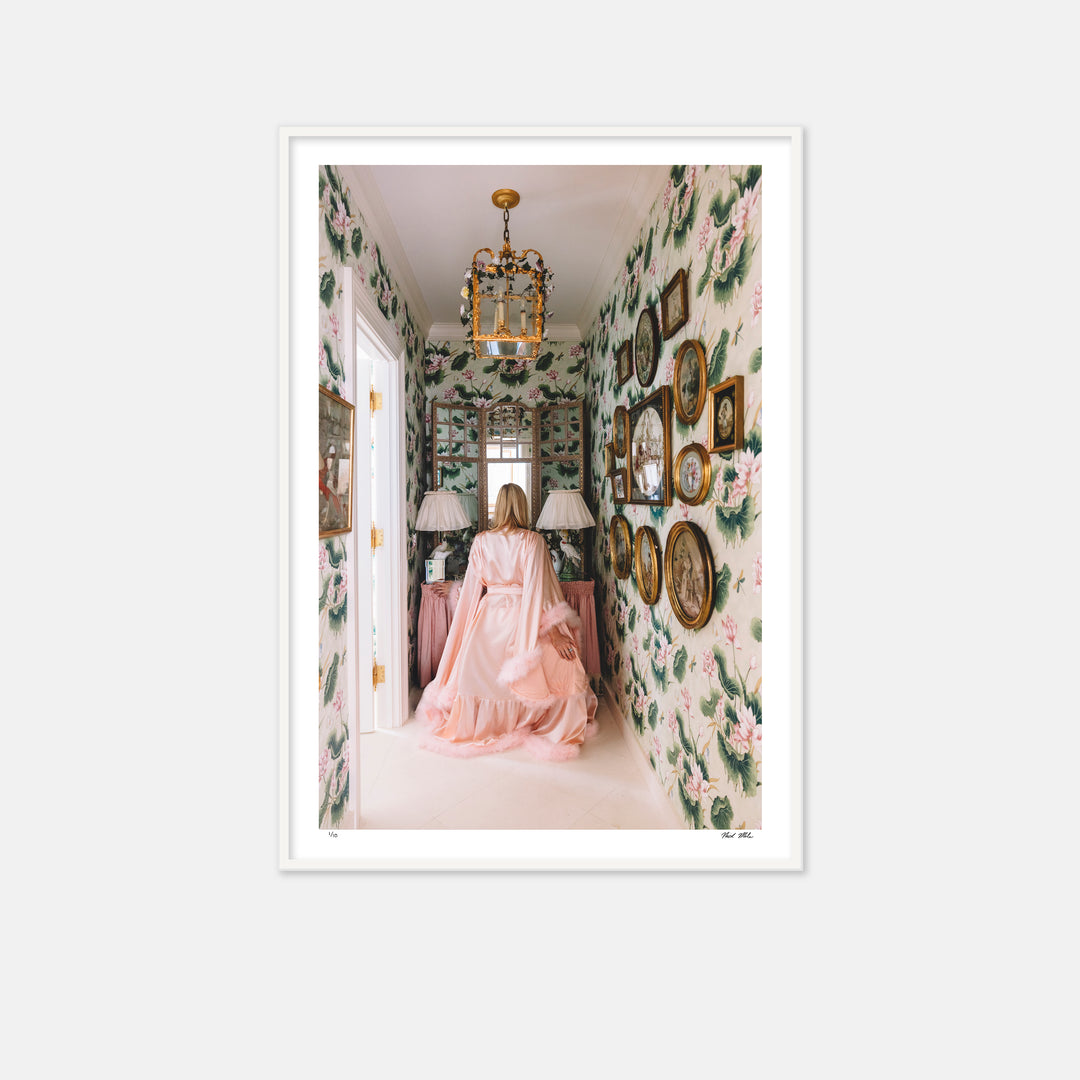 woman sitting with her back to the camera in a pink feather robe staring into a mirror in a room with large floral pink wallpaper.