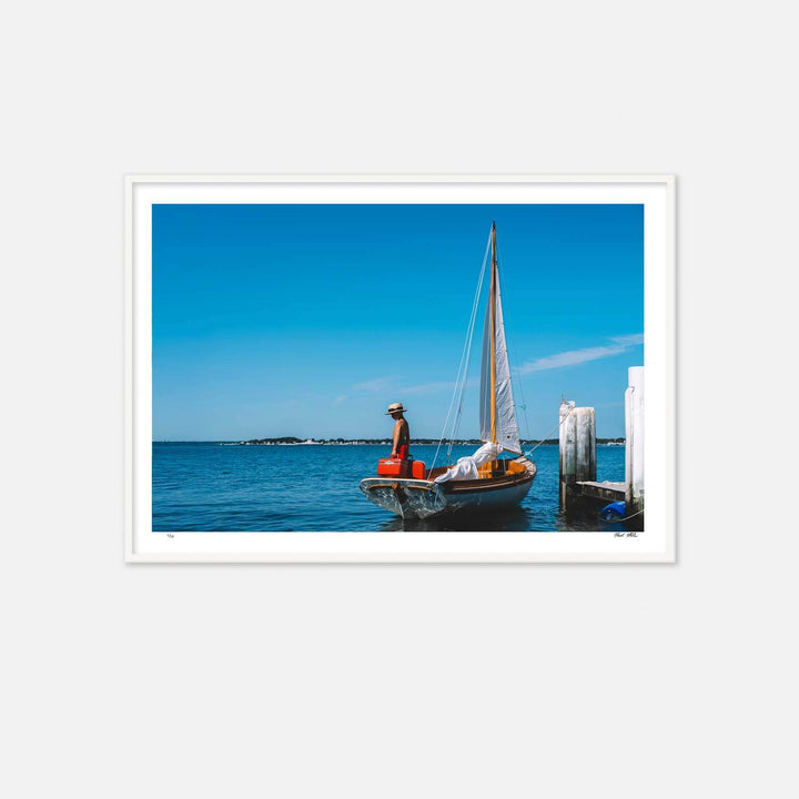 Nick Mele Print, titled Sailing Away. A sailboat with a boy in the boat that is tied to a  dock against a clear blue sky.