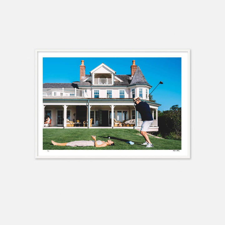 Nick Mele Print, titled Teeing Off. A man playing golf on a well-manicured lawn with a large house in the background. A lady in a dress is laying on the lawn and she has a golf tee in her mouth with a golf ball on the tee.
