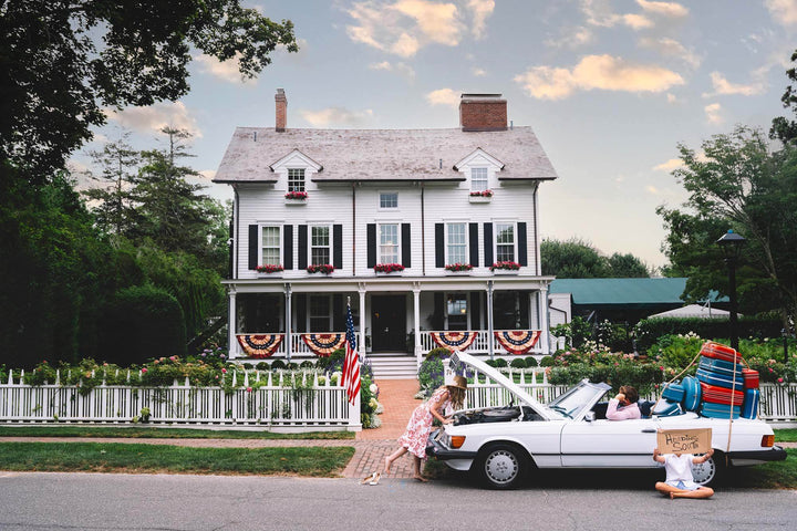Nick Mele Print titled "Heading south". A white house with American flags, a man sitting ind river seat of convertable. Tons of lugage strapped to back of car. A women is leaning into hood of car trying to fixe it and a little boy is sitting outside car with a sign that says Heading south.