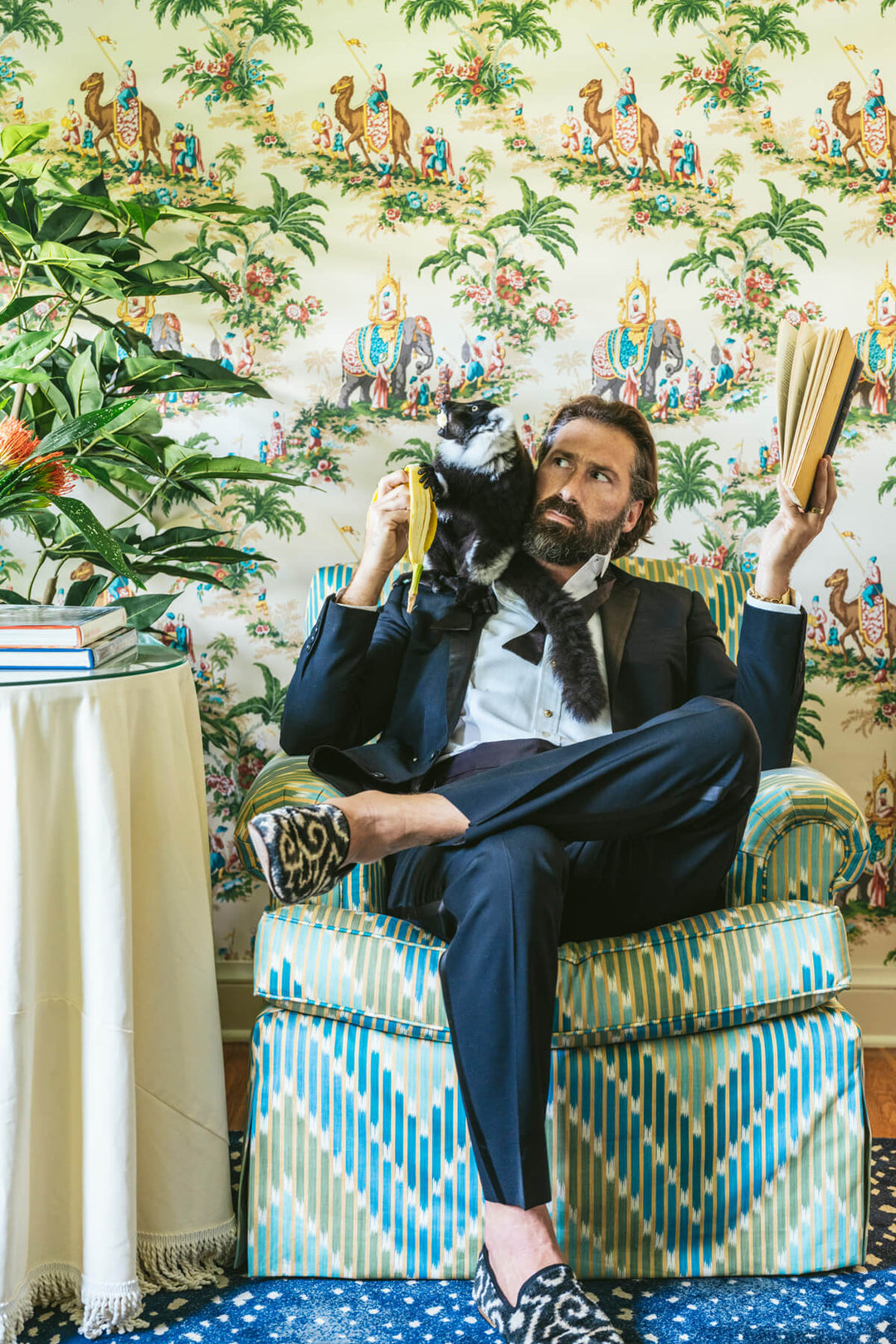 Man in a suit sitting on a patterned chair holding a cat, with decorative wallpaper and plants in the background.