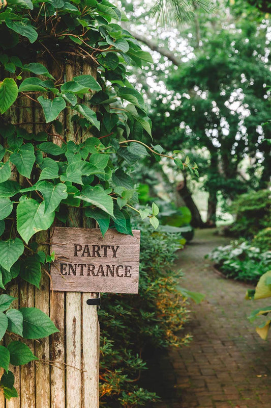 Nick Mele print, titled Party Entrance. A wooden sign pointing to 'Party Entrance' in a lush garden setting
