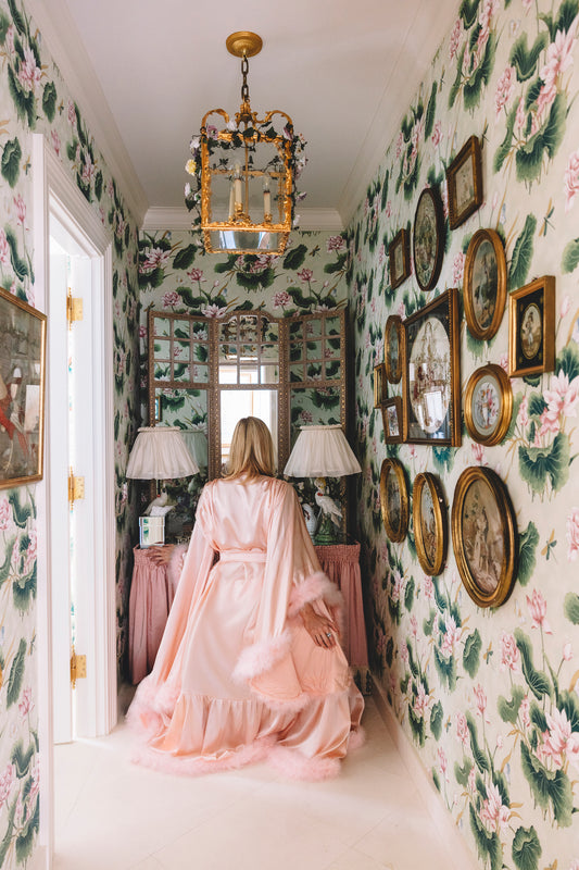 woman sitting with her back to the camera in a pink feather robe staring into a mirror in a room with large floral pink wallpaper.