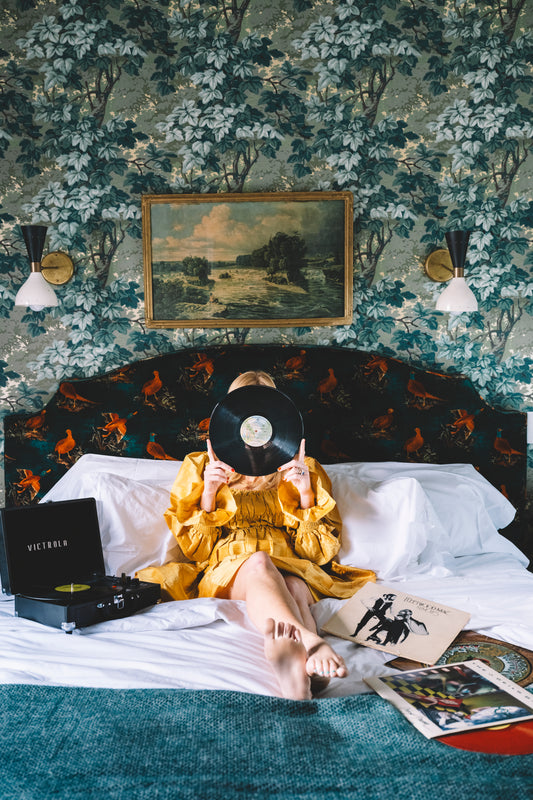 image of girl in yellow dress laying on bed with green leaf wallpaper surrounded by a vintage record player and records. 