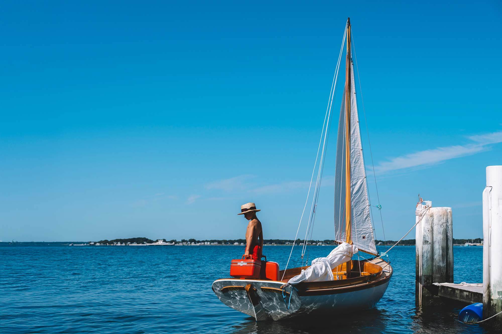 Nick Mele Print, titled Sailing Away. A sailboat with a boy in the boat that is tied to a  dock against a clear blue sky.