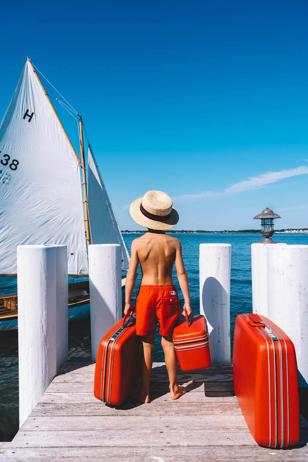 Nick Mele Print titled "Day Trip" - A little boy with red swimsuit and suitcases on a dock with a sailboat in the background