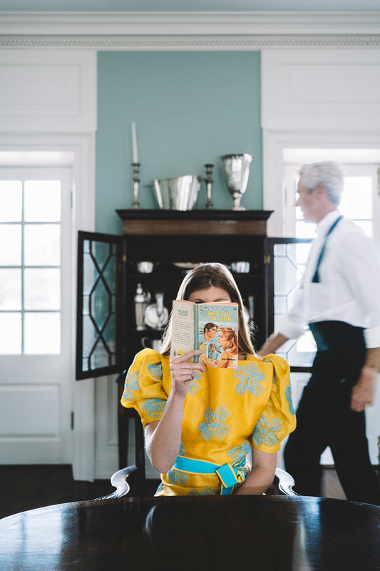 girl reading romance novel with man walking behind to china cabinet