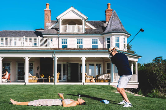 Nick Mele Print, titled Teeing Off. A man playing golf on a well-manicured lawn with a large house in the background. A lady in a dress is laying on the lawn and she has a golf tee in her mouth with a golf ball on the tee.