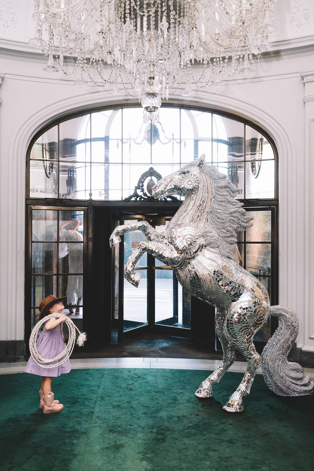 Little cowgirl with a lasso faces a rearing mirrored horse under a crystal chandelier. Nick Mele x Bergdorf Goodman