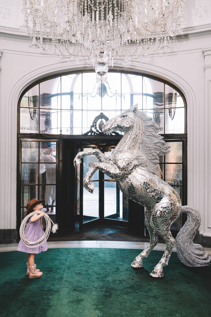 Little cowgirl with a lasso faces a rearing mirrored horse under a crystal chandelier. Nick Mele x Bergdorf Goodman