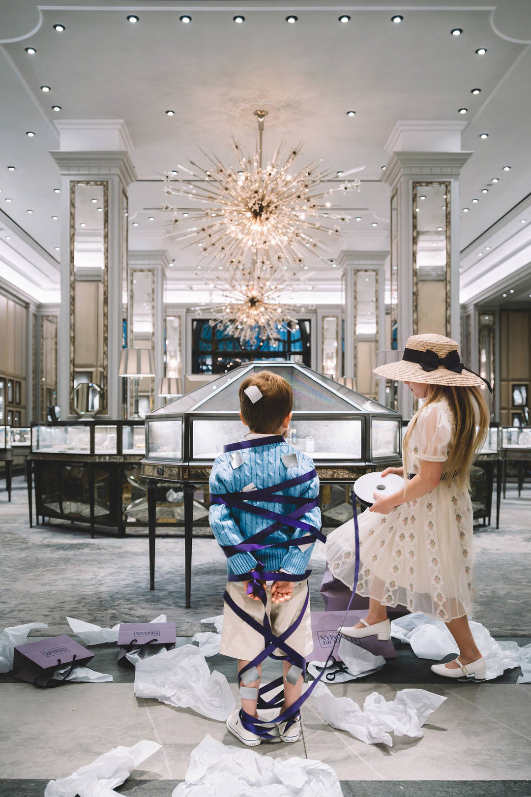 Girl wraps a boy in purple ribbon amid tissue paper in a jewelry salon. Nick Mele x Bergdorf Goodman, A New York Affair