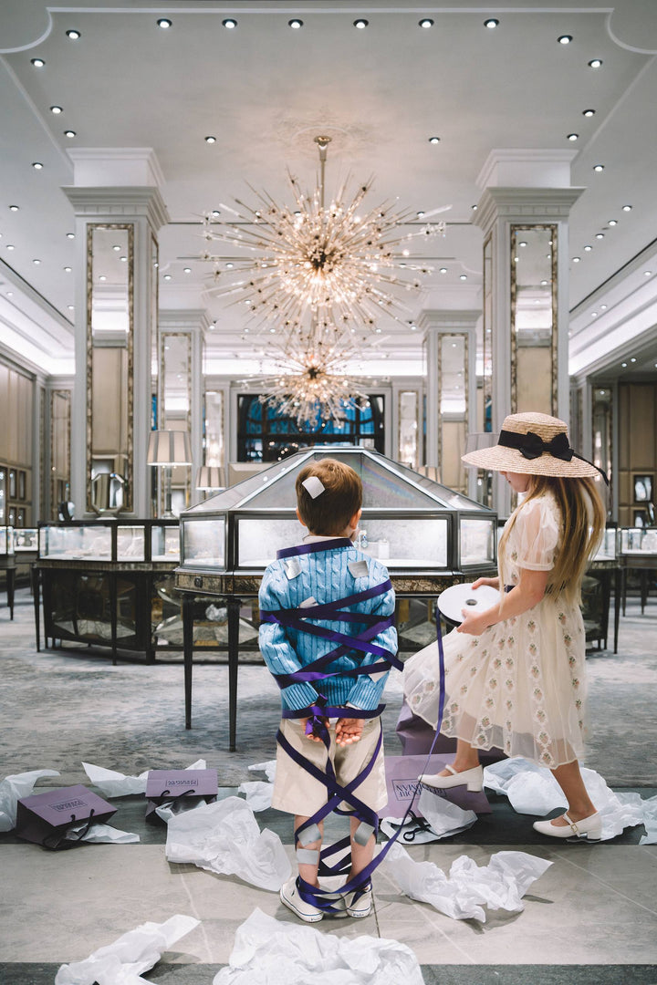 Girl wraps a boy in purple ribbon amid tissue paper in a jewelry salon. Nick Mele x Bergdorf Goodman, A New York Affair