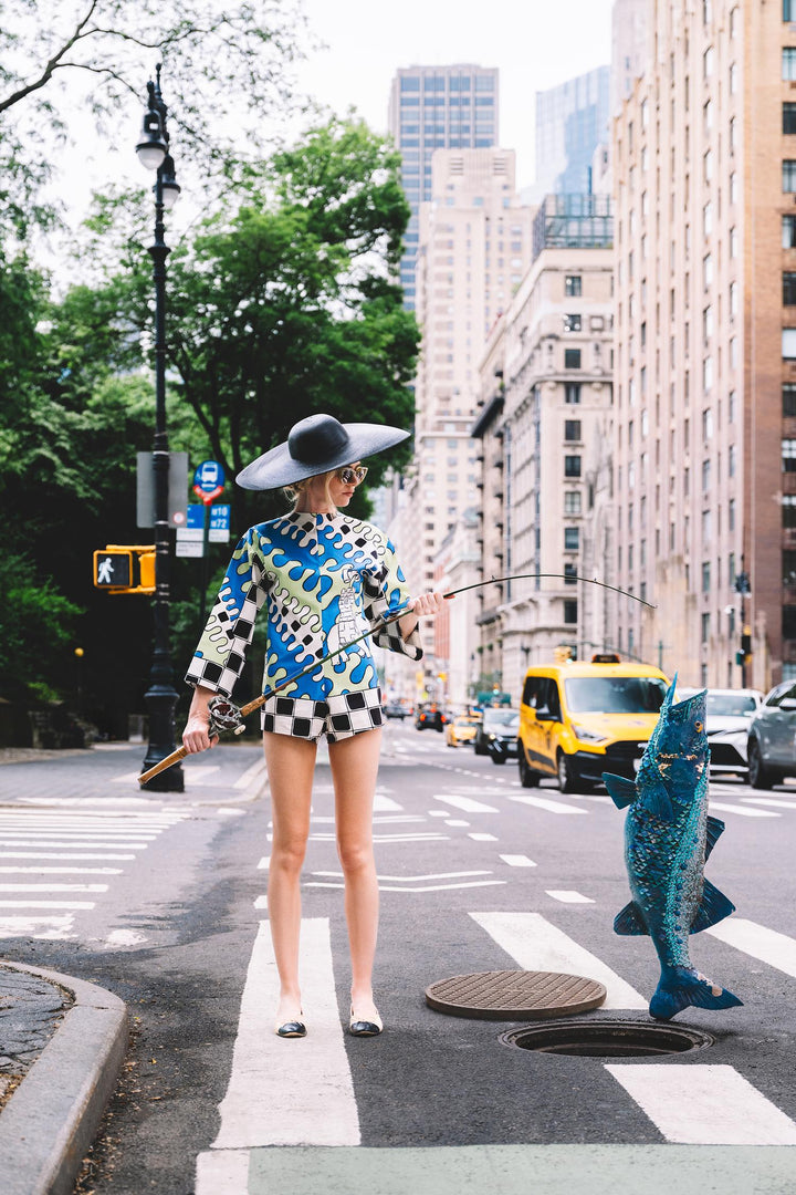 Woman in a wide hat fishes a giant blue fish from a manhole on a New York crosswalk. Nick Mele x Bergdorf Goodman