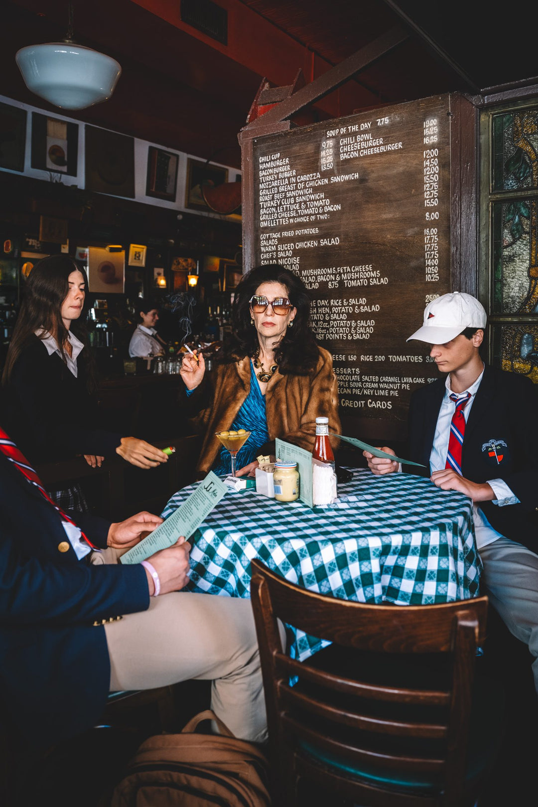Woman in fur holds court with preppy teens at a gingham table in J.G. Melon. Nick Mele x Bergdorf Goodman, A New York Affair