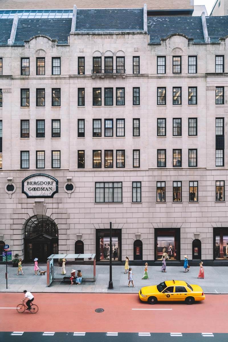 A high-angle street view of a large beige building with many windows, a Bergdolt-Gordmans sign above the entrance, a red street, and a yellow taxi in the foreground.