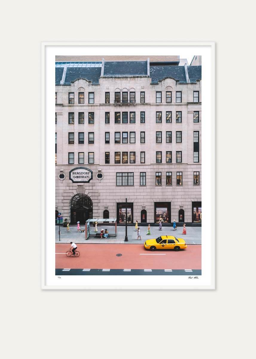 A framed art print featuring a city street scene with a large beige building, a yellow taxi, and a cyclist, displayed against a light beige background.