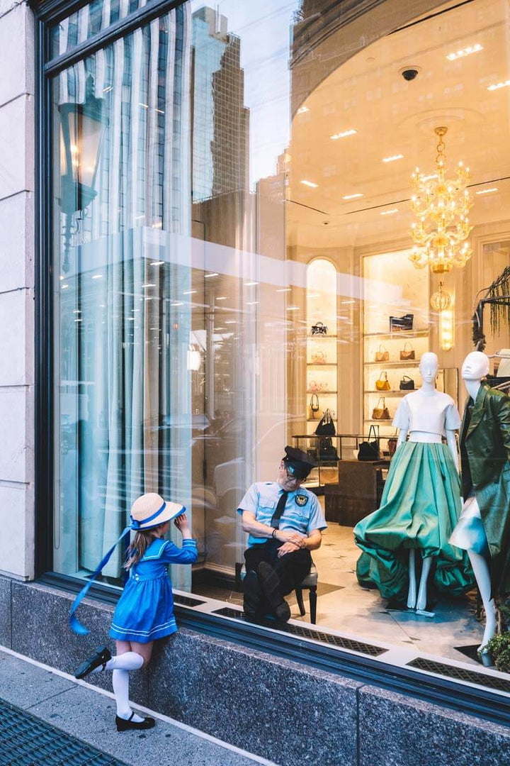 Nick Mele's Night Watch: girl in a blue sailor dress peers into a Bergdorf Goodman window at a sleeping guard