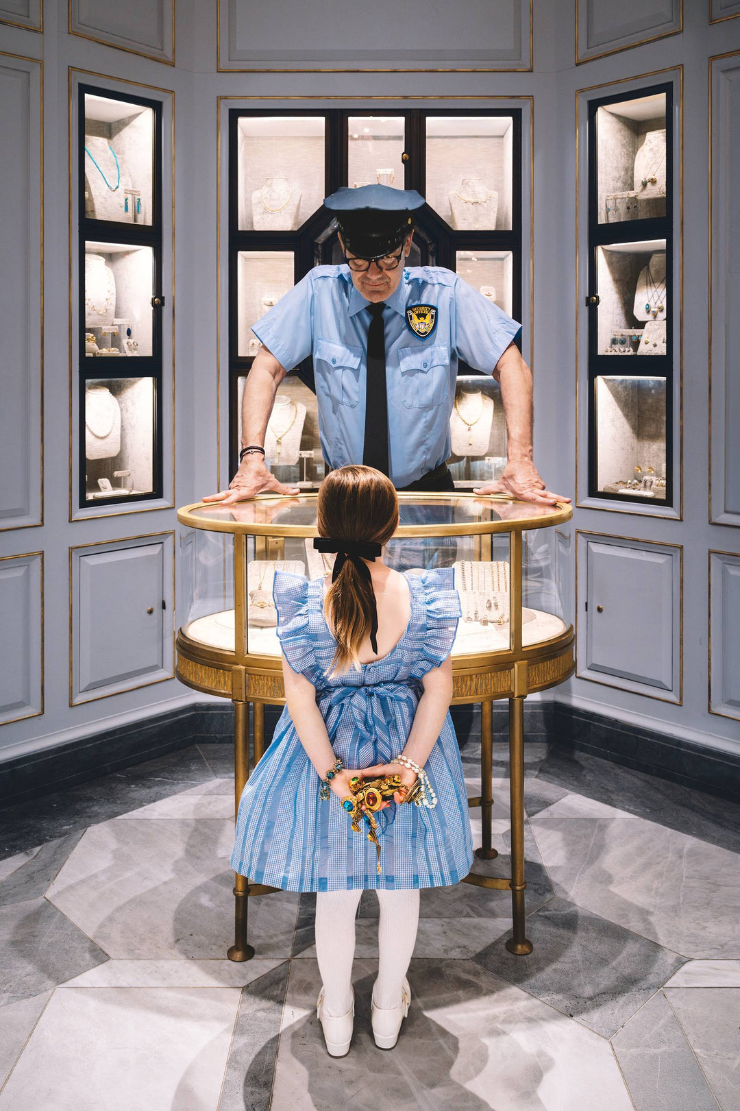 Girl hides jewels behind her back as a guard leans over a jewelry case. Nick Mele x Bergdorf Goodman, A New York Affair