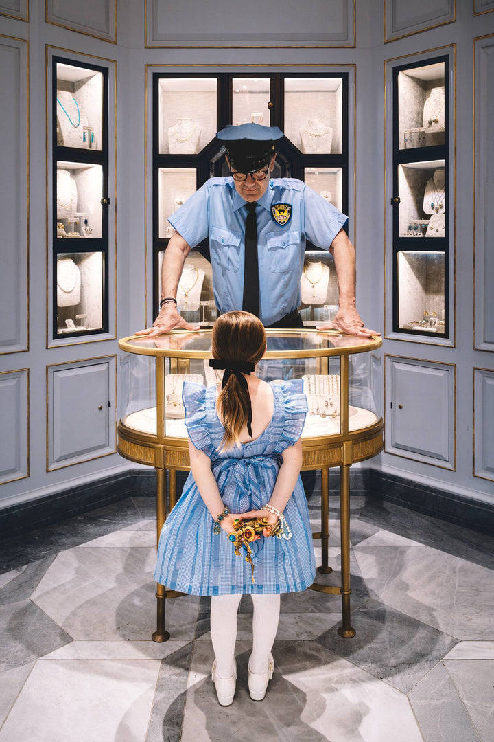 Girl hides jewels behind her back as a guard leans over a jewelry case. Nick Mele x Bergdorf Goodman, A New York Affair